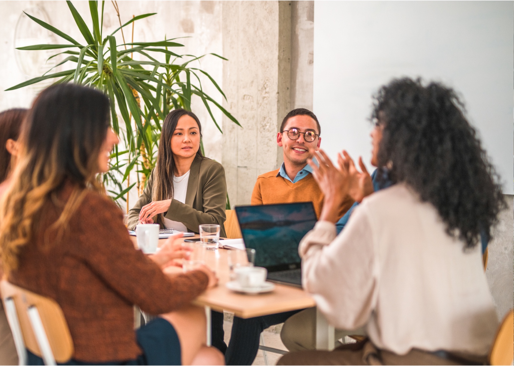 Diverse team collaborates around a table with laptops and drinks, engaging in discussion.