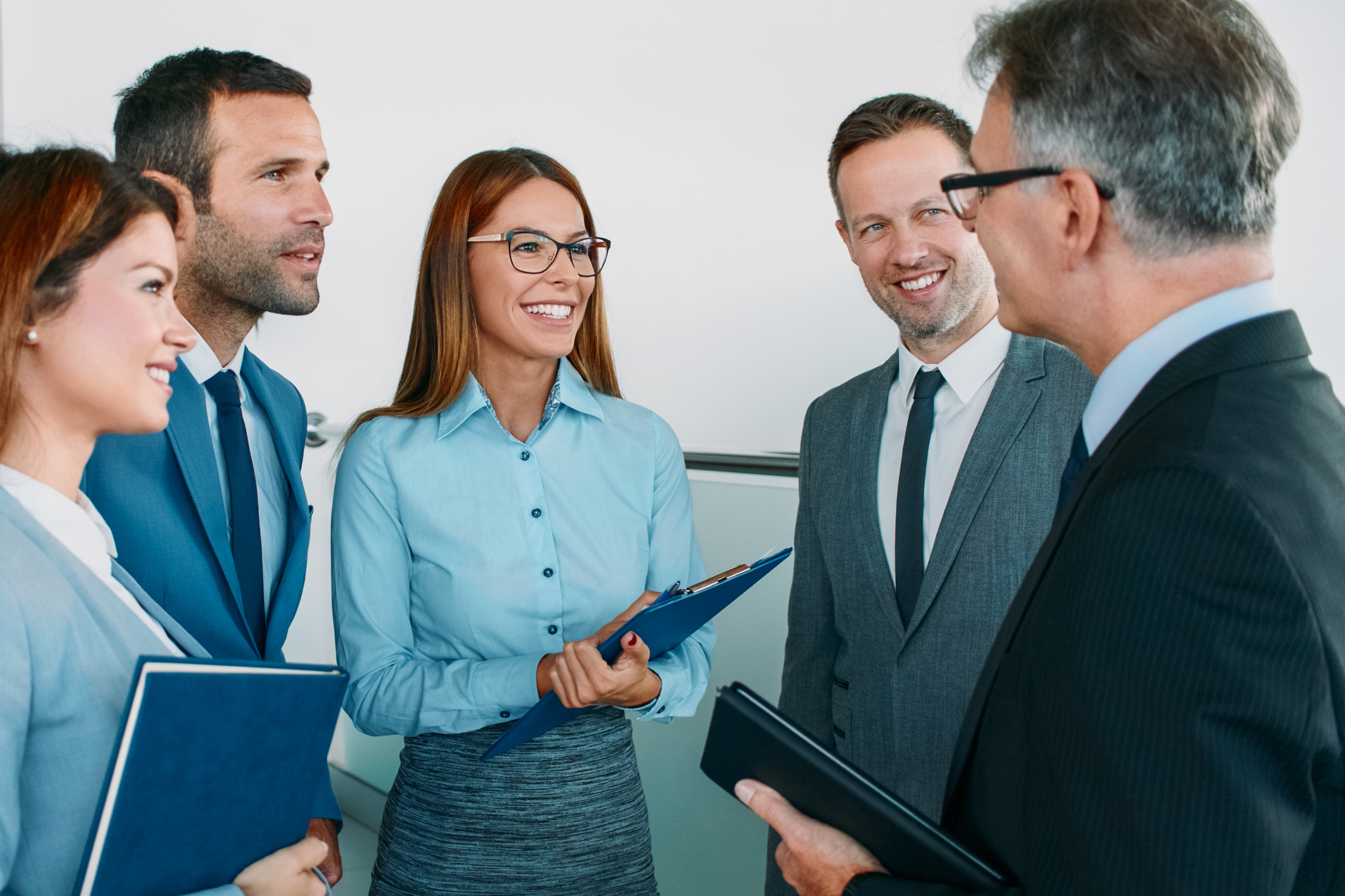 Diverse business team in professional attire smiling and discussing while holding folders and clipboards in a bright office.