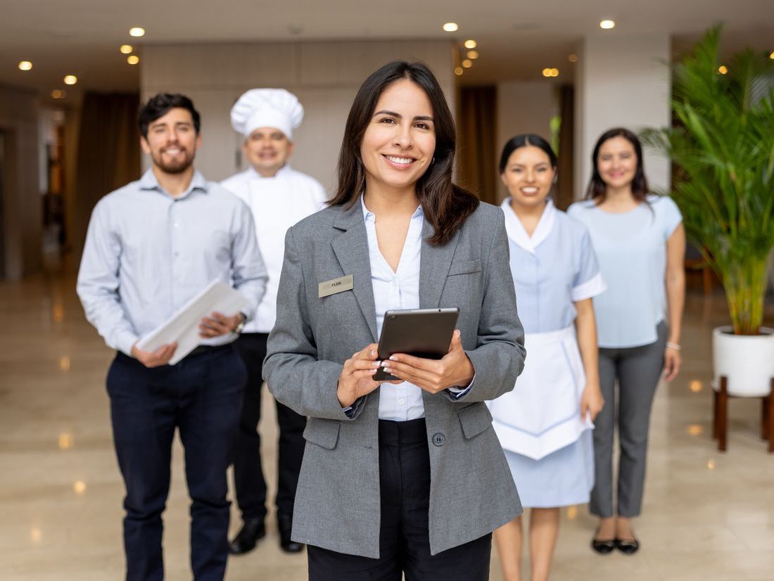 Professional woman in grey blazer holding tablet, smiling, with diverse hospitality staff behind her in a bright lobby.